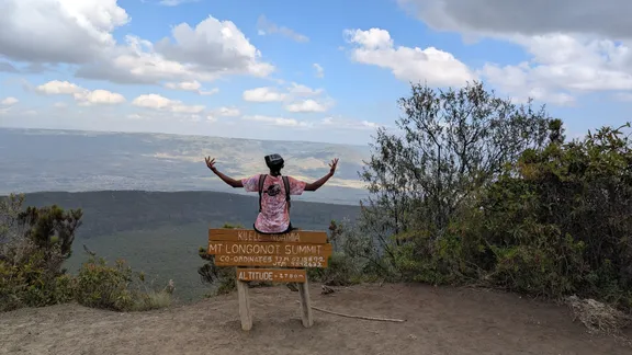 Lewis sitting on the Mt Longonot summit sign with his arms spread over the Rift Valley